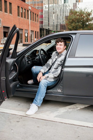 senior boy sits in drivers seat of his car with door open smiling on the streets of downtown OKC
