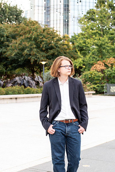 senior boy in a sport coat and jeans walks along a sidewalk looking off to the side with hands in pockets at Myriad Gardens with Devon Tower behind him in Oklahoma City