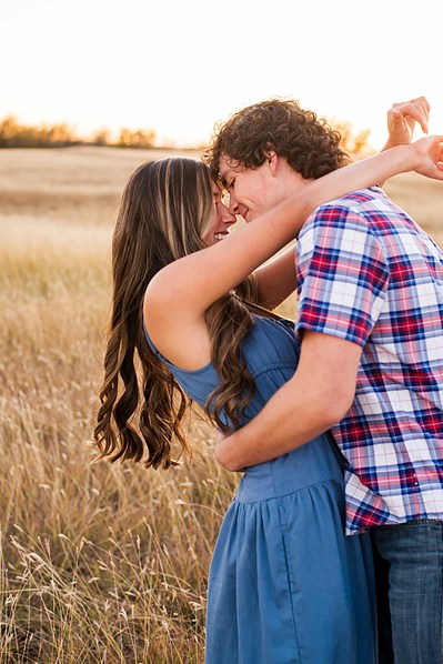 senior girl in a blue dress hugs the neck of her boyfriend in a plaid shirt in a country field in Tuttle Oklahoma