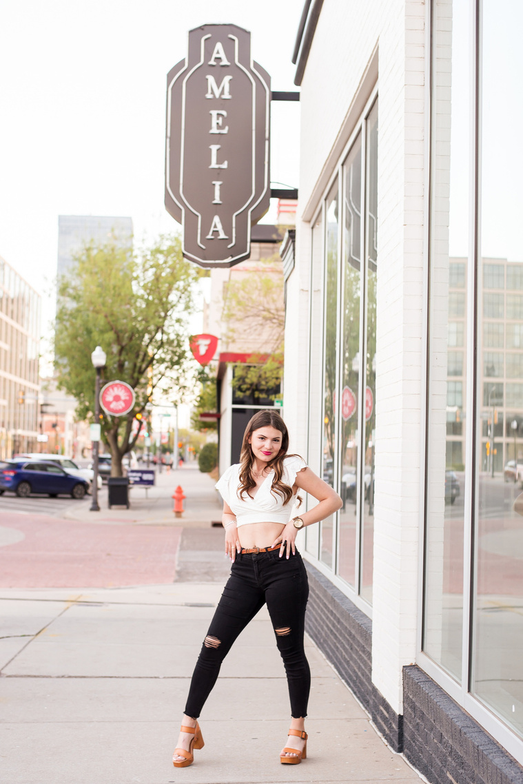 high school senior girl with long brown hair wearing a white crop top, black jeans and heels standing with her hands on her hips on the sidewalk in Automobile Alley in Oklahoma City, Oklahoma