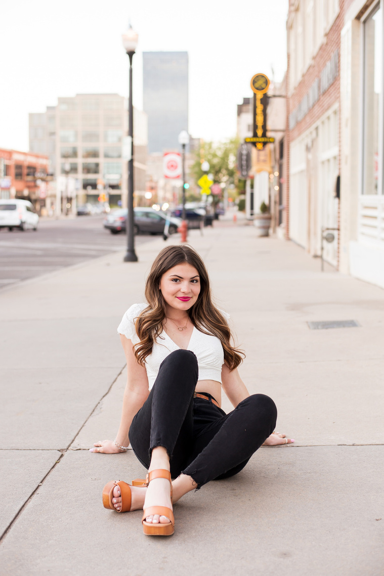 high school senior girl with long brown hair wearing a white top and black jeans sits cross legged on the sidewalk in front of buildings in Automobile Alley in Oklahoma City, Oklahoma
