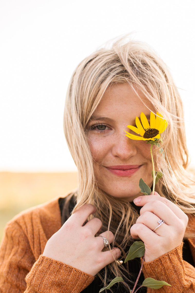 high school senior girl with blond hair wearing a rust colored sweater holding a yellow flower covering one eye, and smiling softly in a field in Oklahoma for her photo session