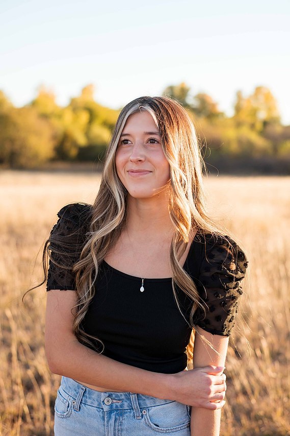 senior girl with long blond hair wearing a black shirt and jeans stands casually and smiles in a field with fall trees behind her in Oklahoma