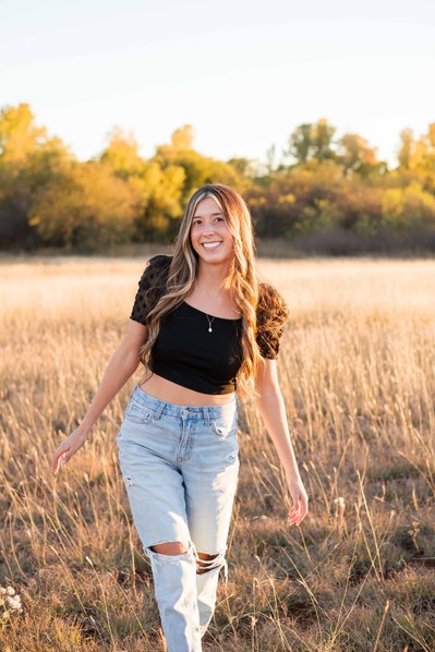 senior girl in a black top and jeans walks in a field with golden grass and trees smiling in Tuttle Oklahoma