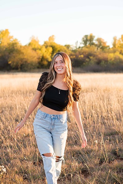 senior girl in a black top and jeans walks in a field with golden grass and trees smiling in Tuttle Oklahoma