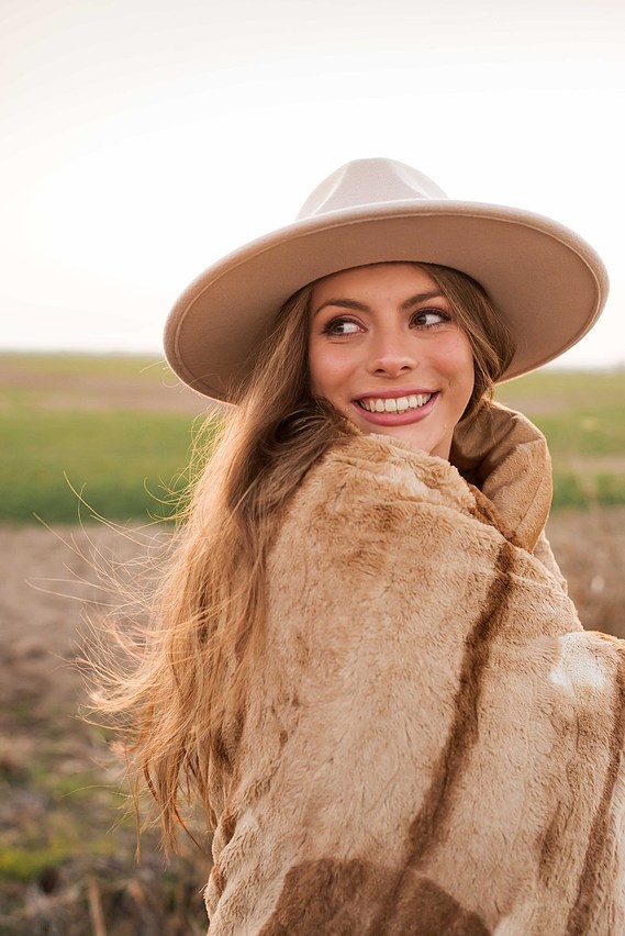 beautiful senior girl wearing a country hat, wrapped in a blanket smiles over her shoulder standing in a field in Oklahoma