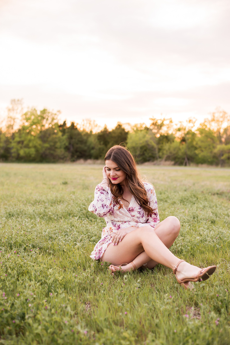 high school senior girl dressed in boho style sits crossed legs looking down and tucking her long hair behind her ear in a field with trees and a sunset behind her in Oklahoma City, Oklahoma