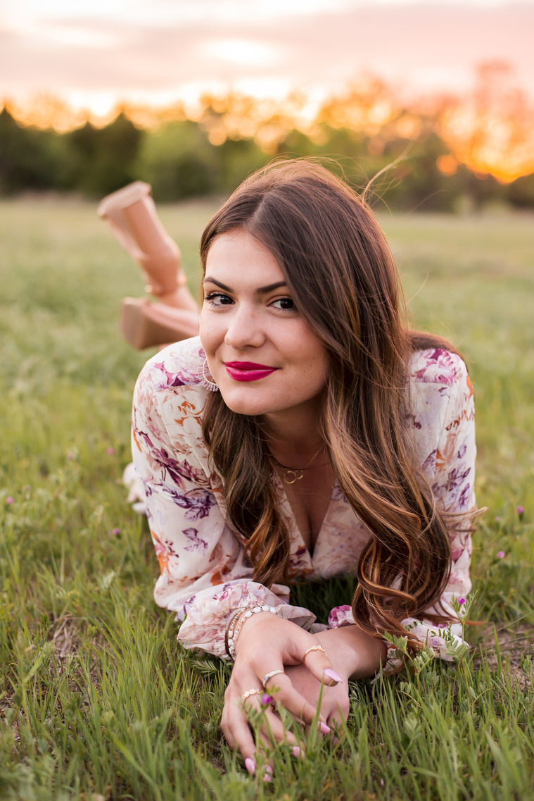 high school senior girl with long brown hair dressed in boho style lays on her stomach and smiles to the side  in a field with trees and a sunset behind her in Oklahoma City, Oklahoma