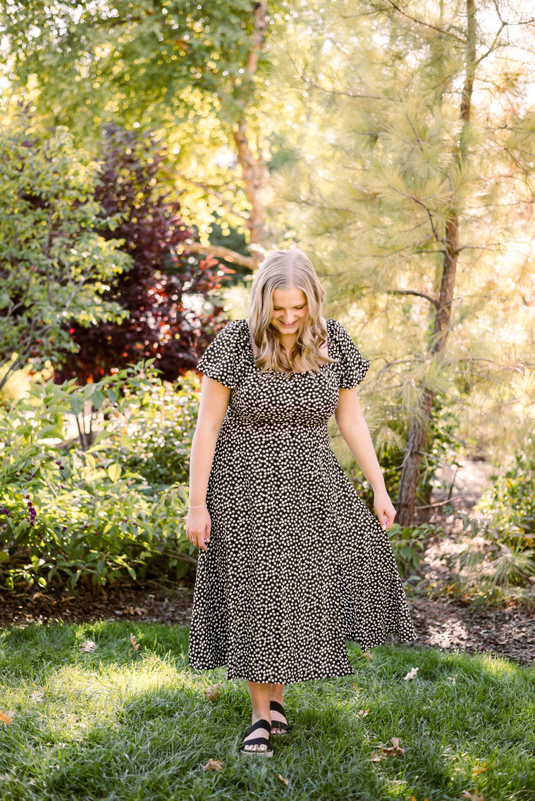 blond high school senior girl wearing a dress walks looking down and smiling in front of trees and plants at Myriad Gardens in downtown Oklahoma City
