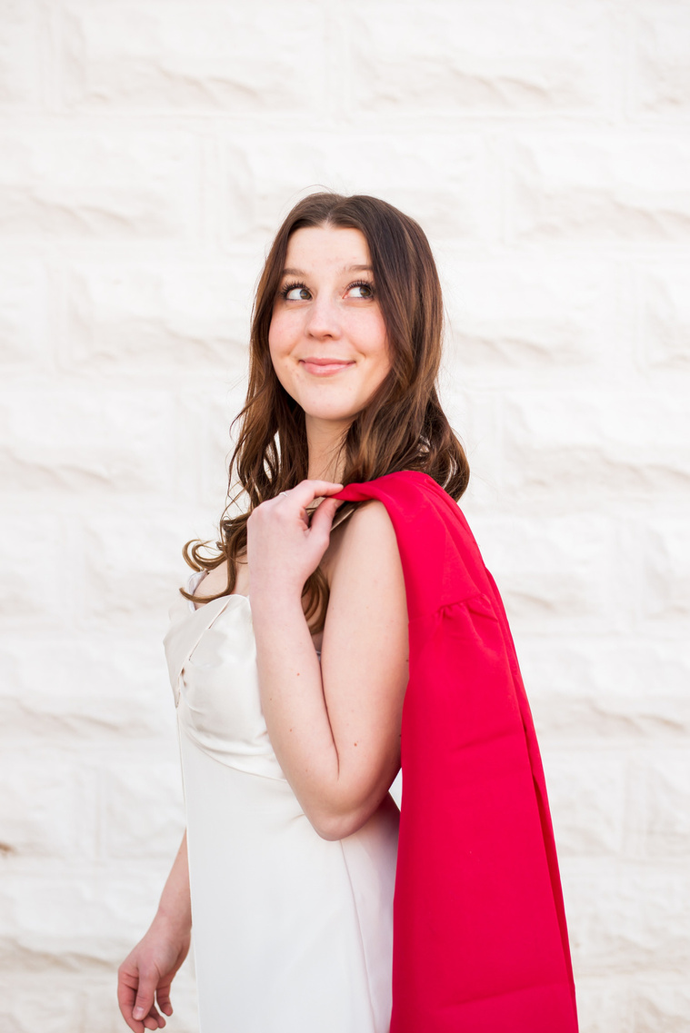 high school senior girl wearing a white dress in front of a white wall holds her red graduation gown over her shoulder and smiles up a the sky in Tuttle Oklahoma