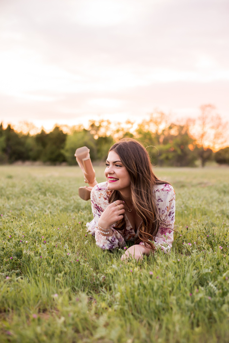 high school senior girl dressed in boho style lays on her stomach and smiles to the side  while playing with her long brown hair in a field with trees and a sunset behind her in Oklahoma City, Oklahoma