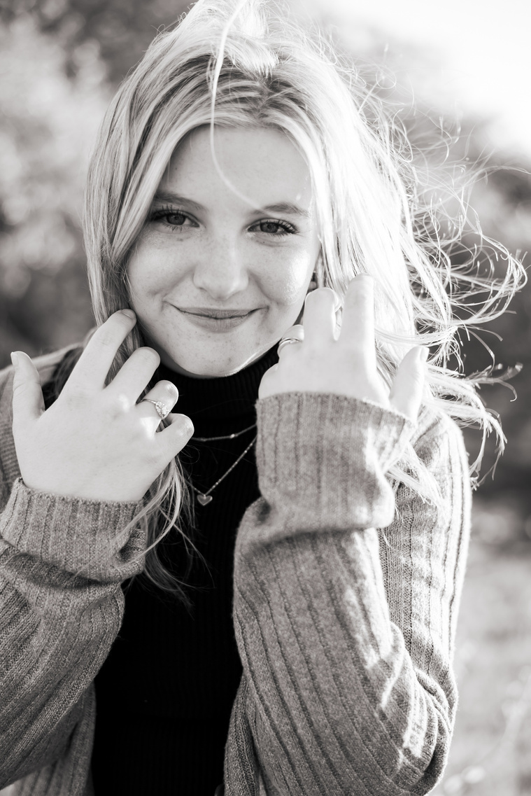 black and white photo of a high school senior girl holding back her hair from her face as the wind blows it to the side, smiling softly