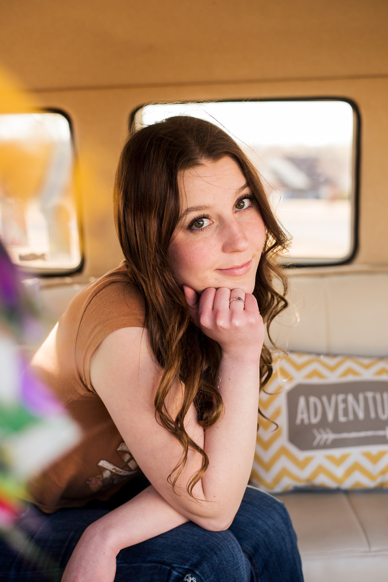 portrait of a high school senior girl with brown hair wearing a t-shirt and bell bottoms sitting with her chin on her hand and softly smiling inside a yellow and white VW bus in a yard in Tuttle Oklahoma