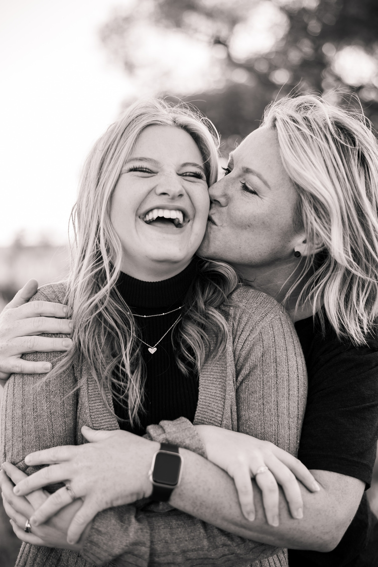 black and white photo of a high school senior girl laughing while her mom hugs her and kisses her cheek
