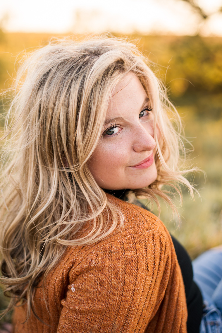 close up photo of a high school senior girl with blond hair wearing a rust colored sweater and jeans, sitting in a golden field looking over her shoulder at the camera
