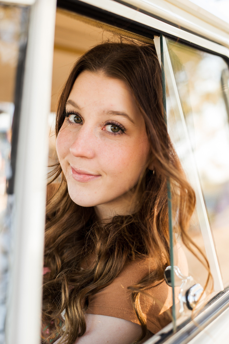 portrait of a high school senior girl with brown hair wearing a t-shirt and bell bottoms sitting in the drivers side of a VW bus in a yard in Tuttle Oklahoma looking out the open window with a soft smile
