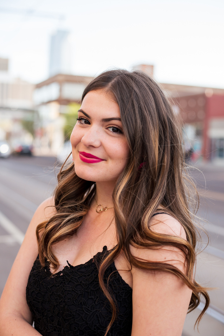 portrait of a high school senior girl with long brown hair in a black dress standing on the sidewalk and smiling softly in Automobile Alley in Oklahoma City, Oklahoma