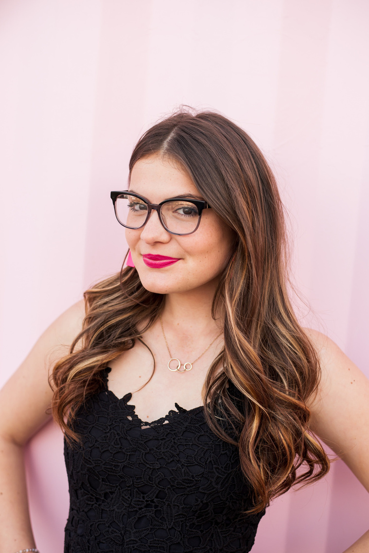 high school senior girl with glasses and long brown hair wearing a black dress and pink earrings standing in front of a pink wall in Automobile Alley in Oklahoma City, Oklahoma