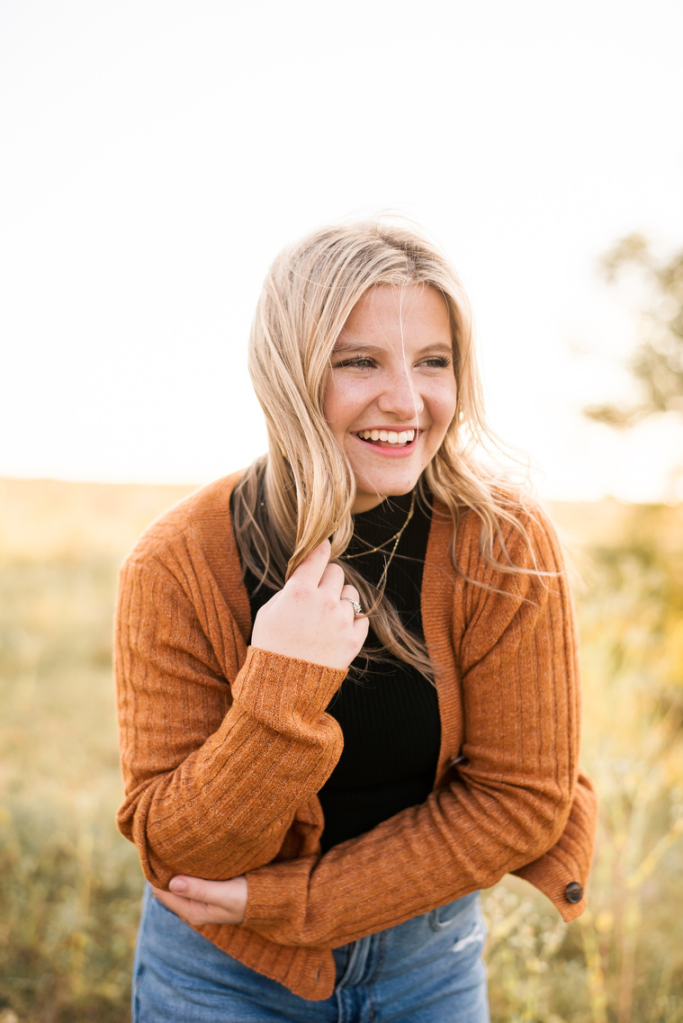 high school senior girl with blond hair wearing a rust colored sweater and jeans,  in a golden field in Oklahoma looking away and laughing for her photo session