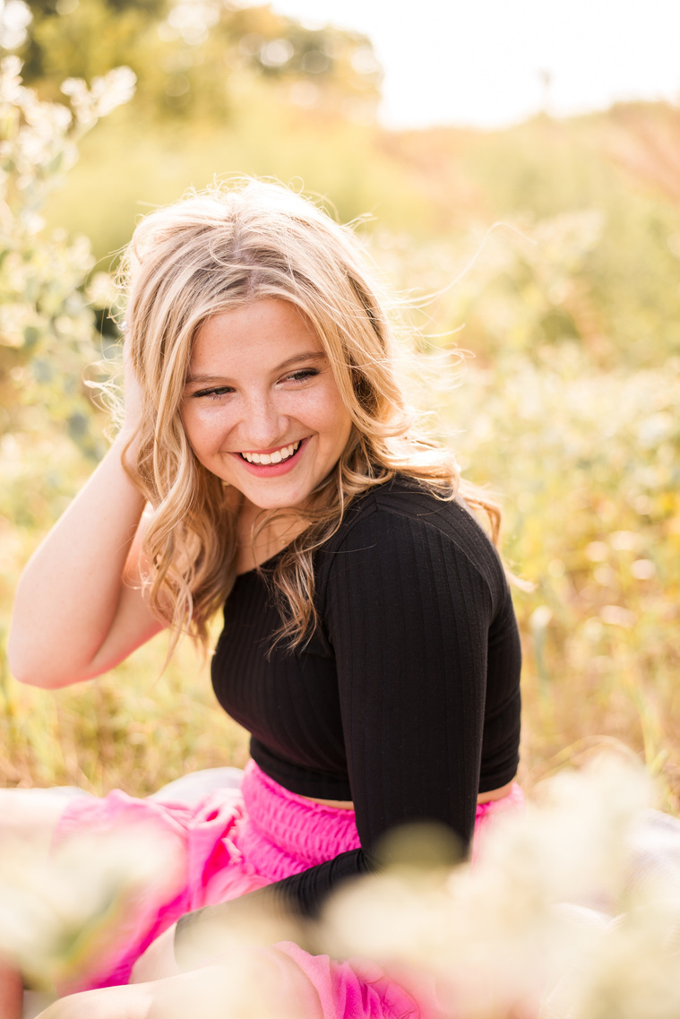 high school senior girl with blond hair wearing a pink skirt and black top, sitting in a golden field in Oklahoma surrounded by milkweed flowers looking down her shoulder with a hand in her hair and smiling for her photo session