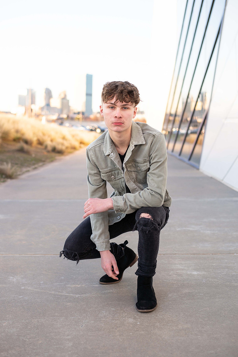 teenage boy wearing a gray jacket and black pants squats on a sidewalk casually next to the Devon Boathouse with the OKC skyline behind him