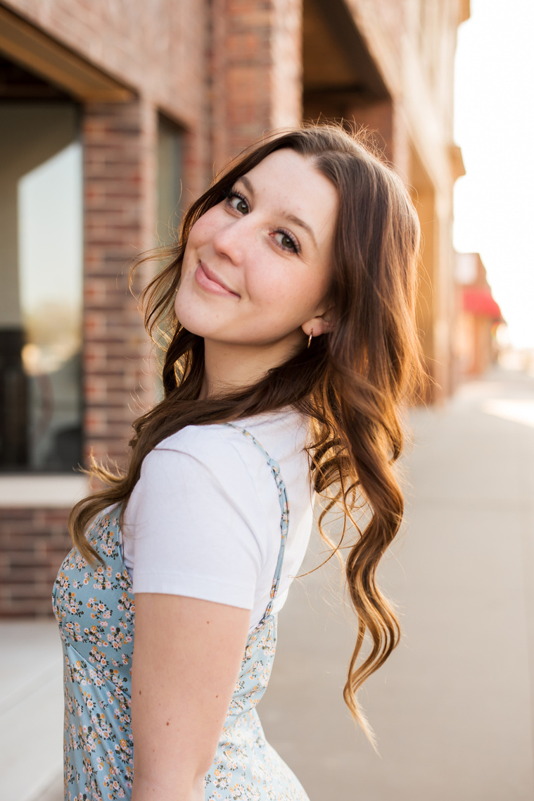 high school senior girl wearing a blue dress stands on the sidewalk smiling over her shoulder with her long brown hair flowing in downtown Tuttle Oklahoma