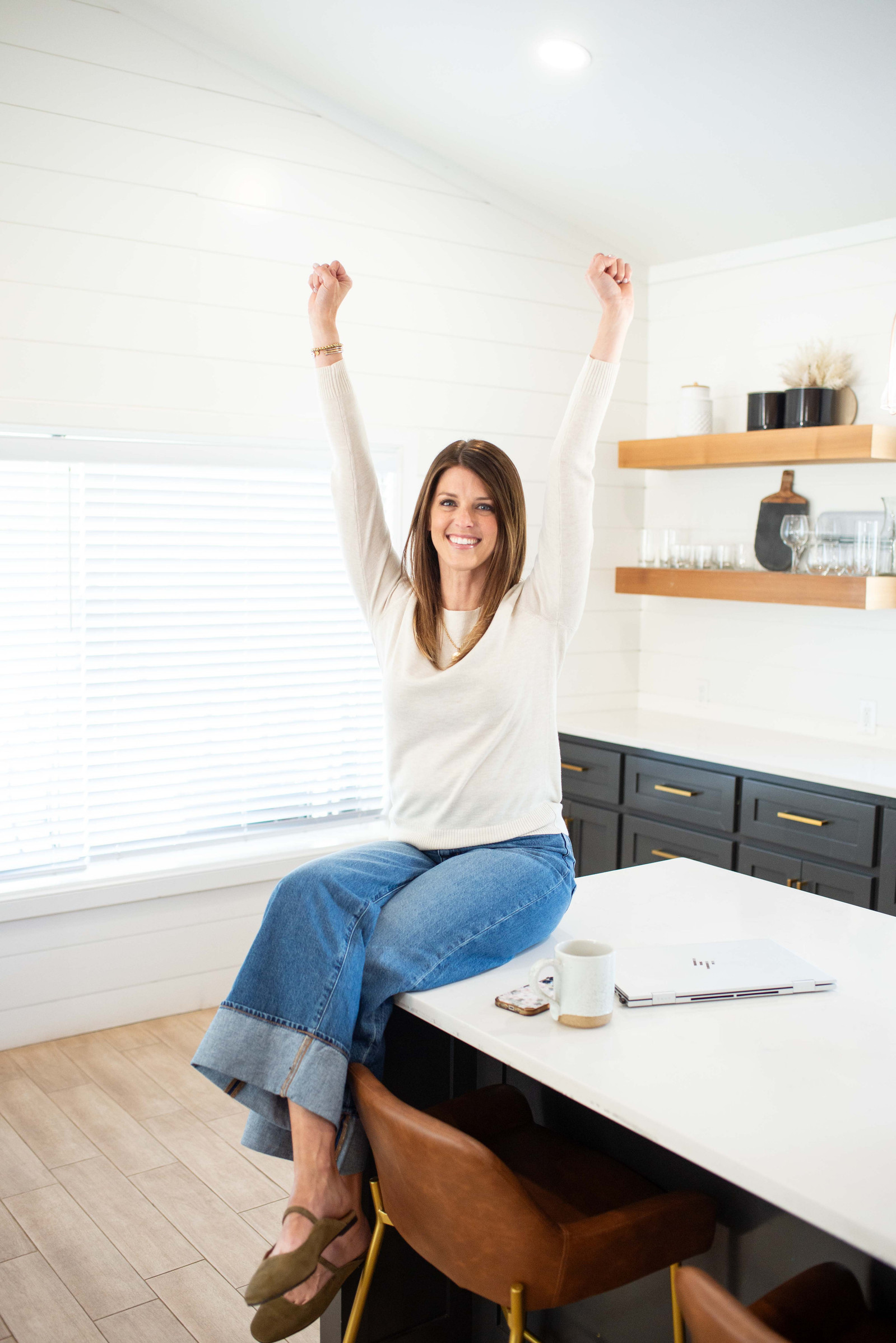 at a personal brand session for a female realtor, she sits on a kitchen island and holds both arms up in a celebration pose and smiles