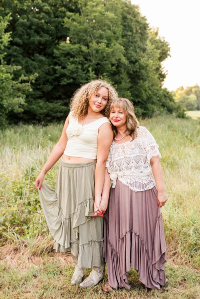a mom and her teen daughter hold hands and stand together smiling in front of trees and a field in Norman