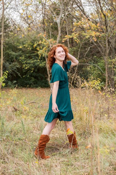 senior girl with red hair wearing a teal dress and boots walks among the grass and trees smiling over her shoulder at Sutton park in Norman Oklahoma