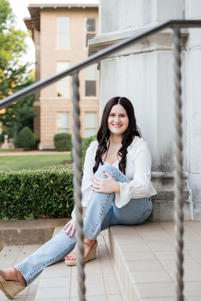 college senior girl in a white top and jeans sits on steps of Chemistry building smiling and hugging one knee on OU campus in Norman