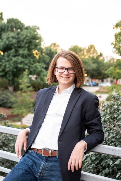 senior boy in a sport coat and jeans leans against a rail smiling with the Myriad Gardens in OKC behind him