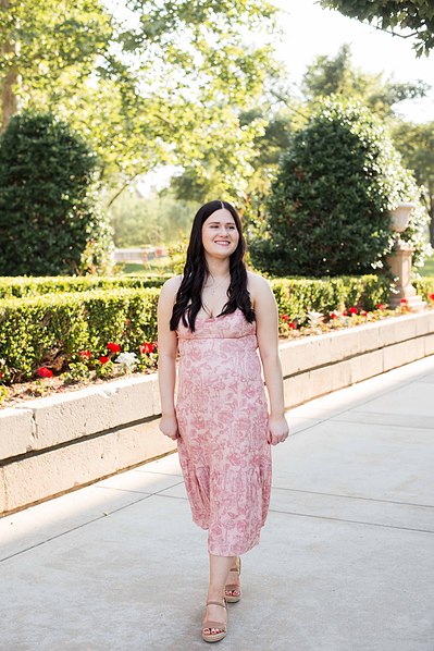 senior girl in a dress walks along a sidewalk with flowers and trees behind her near the Parrington Oval on OU campus in Norman Oklahoma