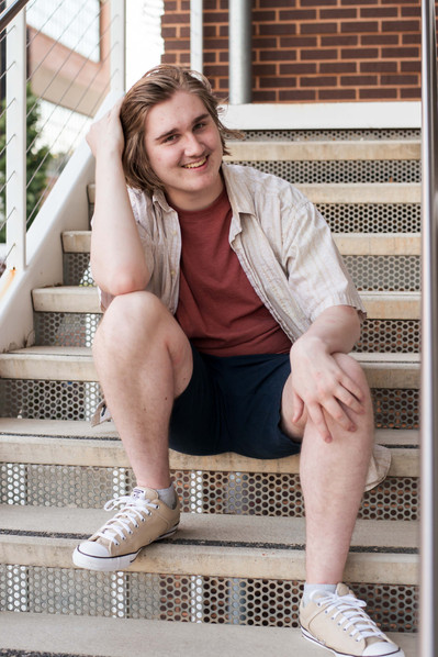 senior boy sits on steps of a parking garage in shorts and tennis shoes, smiling at camera as he brushes hair back with one hand in midtown OKC