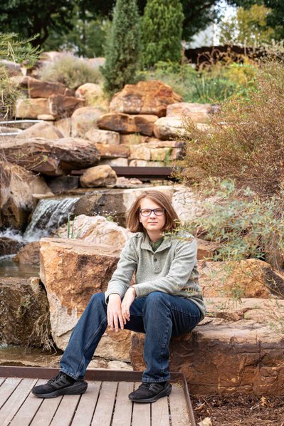 senior boy in green shirt and jeans sits among rocks and a waterfall at Myriad Gardens in downtown Oklahoma City
