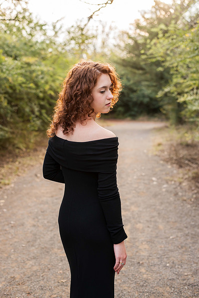 senior girl in a bodycon black dress faces away and looks over her shoulder on a path with woods around it at a park in Norman Oklahoma