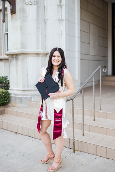 college senior girl in a white dress and stole holds her cap in front of her smiling with the chemistry building of the OU campus behind her