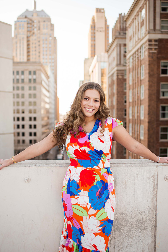 beautiful senior girl wearing a bright floral dress stands with her arms resting on the ledge of the rooftop of a parking garage in downtown Oklahoma City