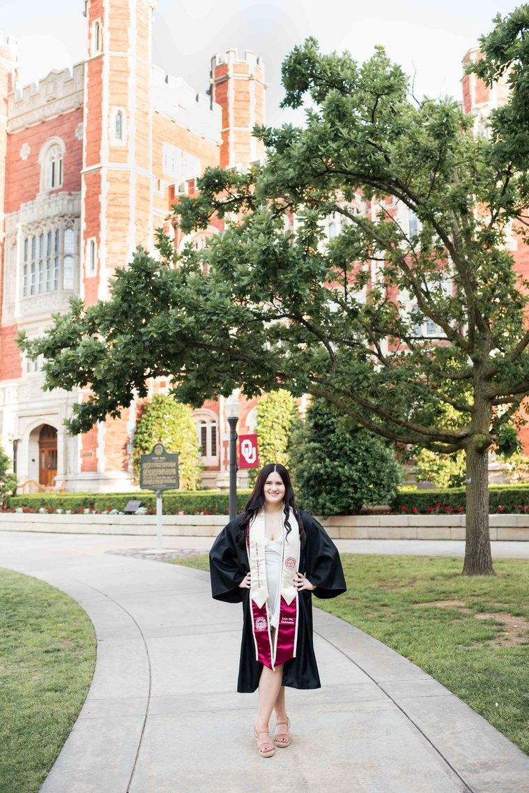 college senior girl wearing a graduation gown and stole stands on a sidewalk on the OU campus in Norman Oklahoma
