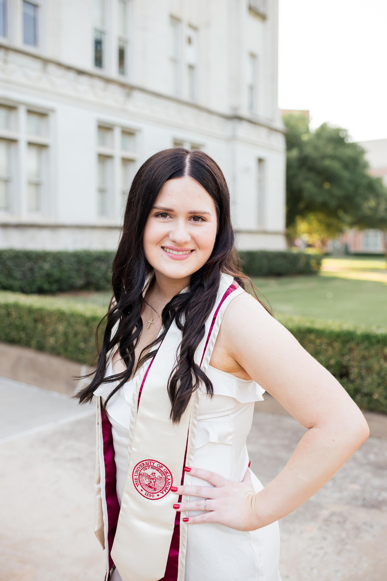 portrait of a college senior with hand on hip smiling wearing a dress and her graduation stole standing in front of the Chemistry building on OU campus in Norman, Oklahoma