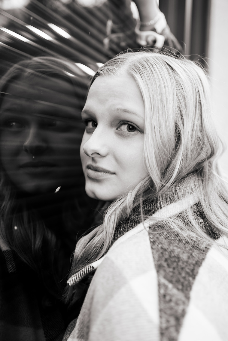 black and white image of a high school senior girl standing against a reflective window and looking at camera with a serious face