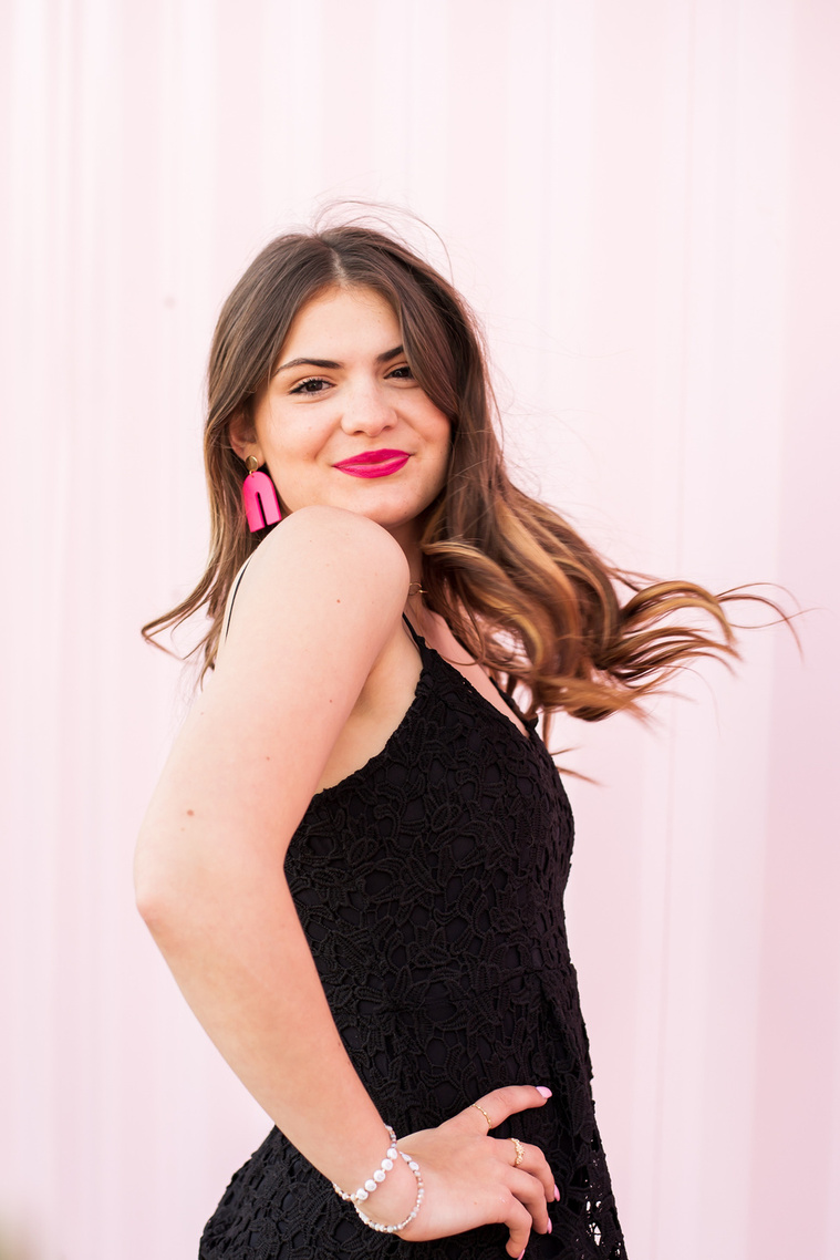 high school senior girl wearing a black dress and pink earrings turns with hair flying toward the camera and smiles in front of a pink wall in Automobile Alley in Oklahoma City, Oklahoma