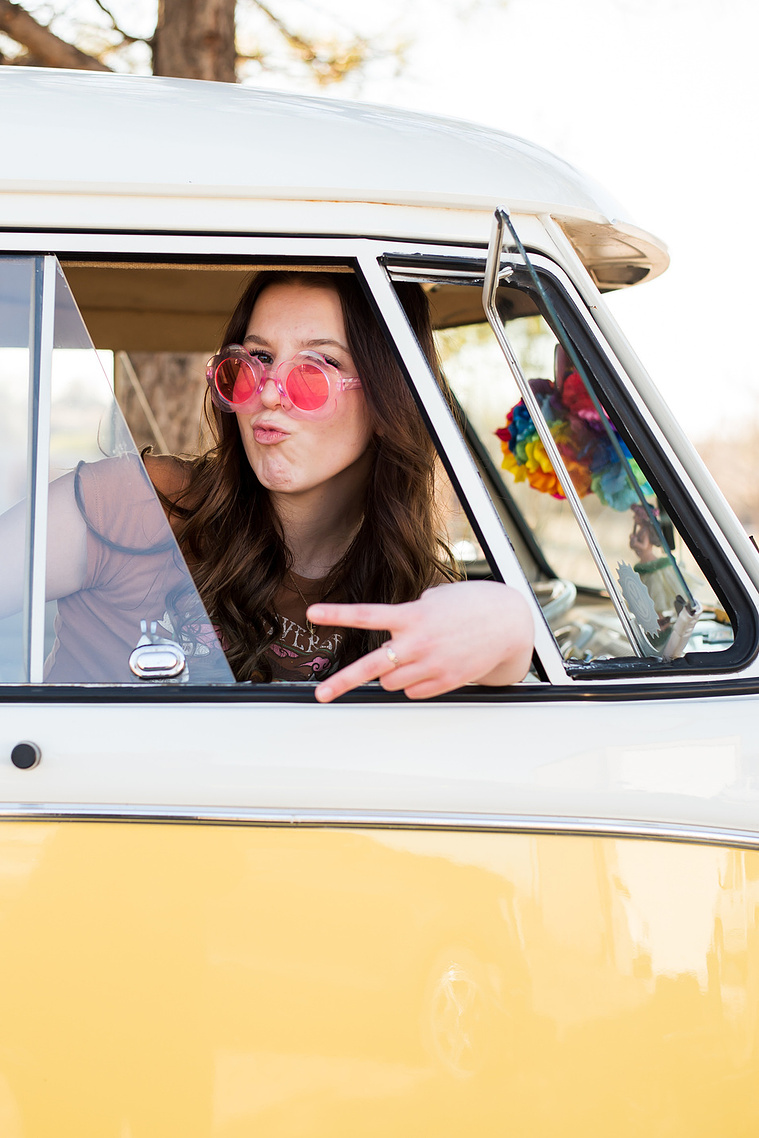 high school senior girl with brown hair wearing a t-shirt and bell bottoms and pink flower sunglasses sits in the passenger side of a yellow and white VW bus in a yard in Tuttle Oklahoma making a peace sign looking out the open window