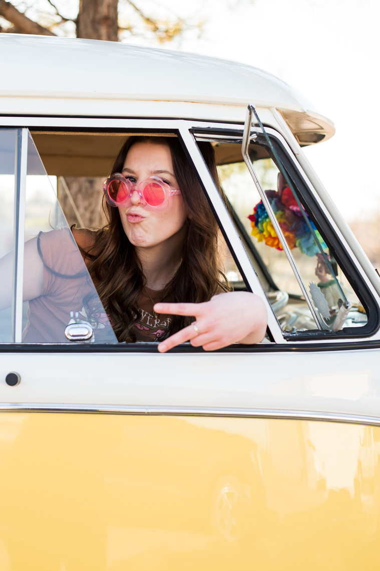 high school senior girl with brown hair wearing a t-shirt and bell bottoms and pink flower sunglasses sits in the passenger side of a yellow and white VW bus in a yard in Tuttle Oklahoma making a peace sign looking out the open window