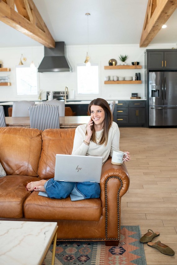 a realtor is working from home sitting on her couch with the kitchen behind her and laptop on her lap, on the phone with a client smiling and holding her coffee mug in a branding photo
