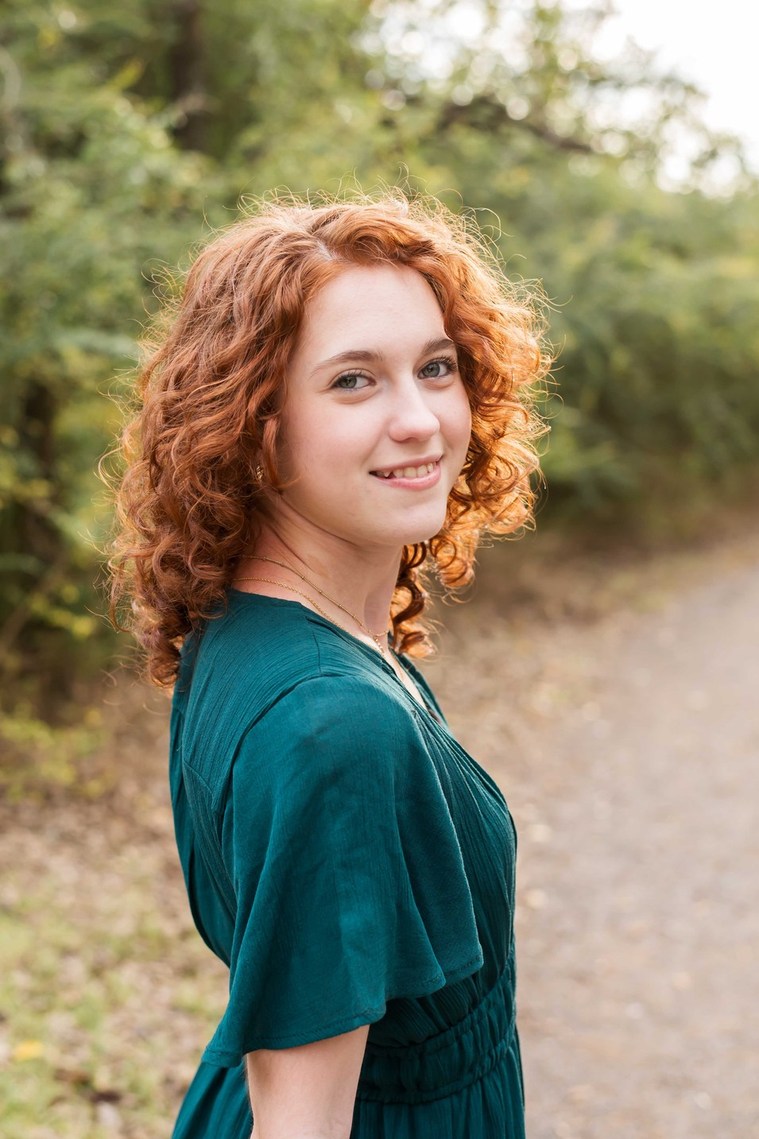 photo of a red headed senior girl wearing a green dress smiling over her shoulder on a wooded path at Sutton Park in Norman, Oklahoma