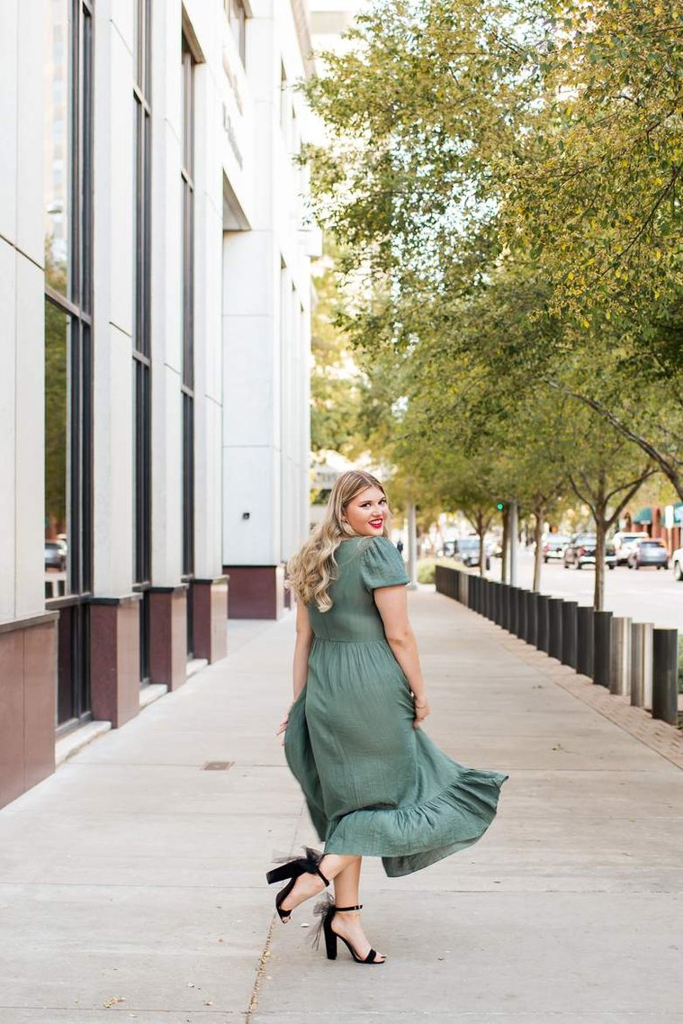 blond high school senior girl in a green dress twirls and kicks her fancy high heel up and smiles on a sidewalk in downtown Oklahoma City for senior portraits
