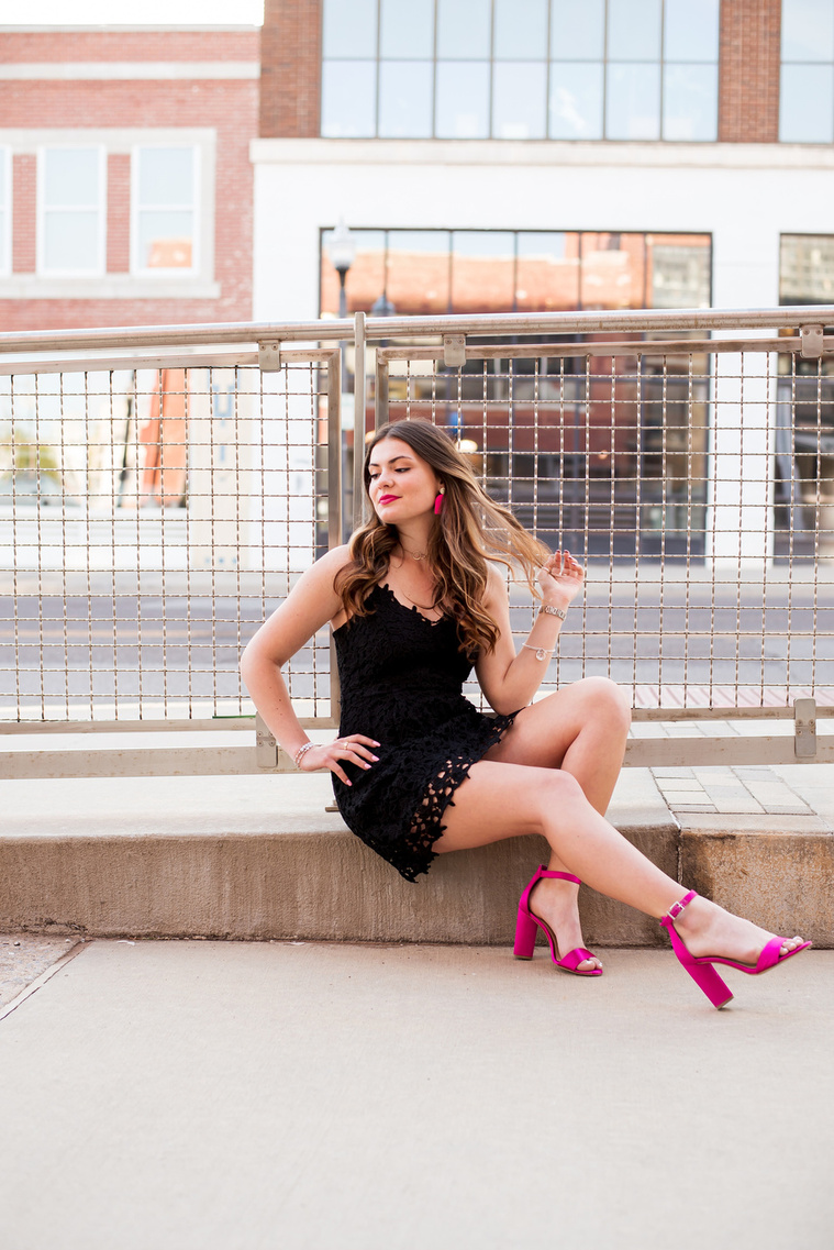 high school senior girl wearing a black dress and pink heels sits on the curb with one hand on her hip and the other in her hair smiling sassy in Automobile Alley in Oklahoma City, Oklahoma