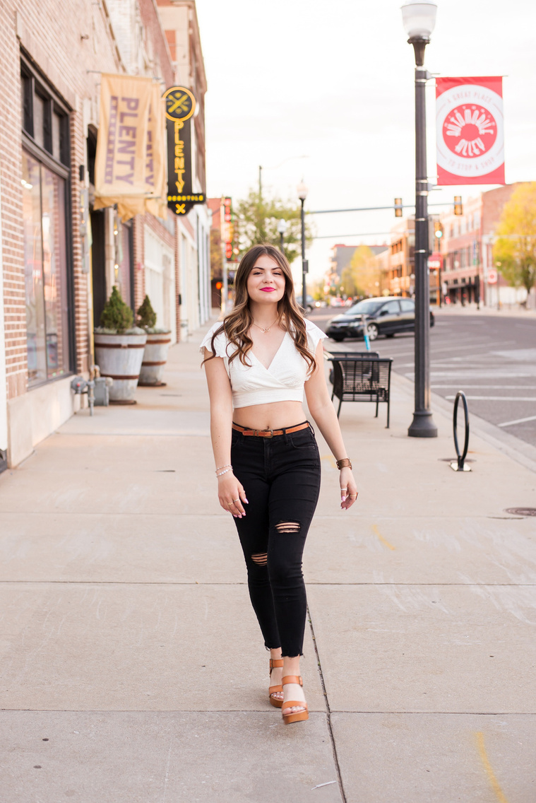 high school senior girl with long brown hair wearing a white top and black jeans walks on the sidewalk in Automobile Alley in Oklahoma City, Oklahoma