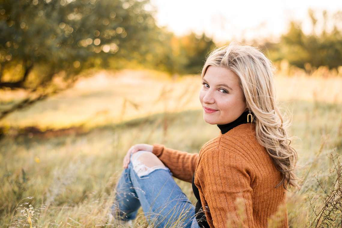 high school senior girl with blond hair wearing a rust colored sweater and jeans, sits in a golden field in Oklahoma looking over her shoulder and smiling for her photo session