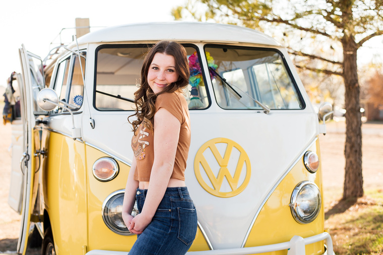 high school senior girl with brown hair wearing a t-shirt and bell bottoms stands to the side and looks over her shoulder in front of a yellow and white VW bus in a yard in Tuttle Oklahoma